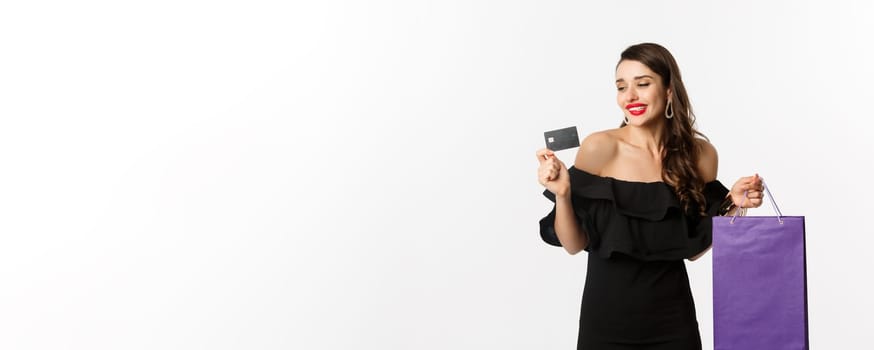 Stylish young woman in black dress going shopping, holding bag and credit card, smiling pleased, standing over white background.