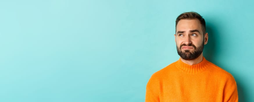 Close-up of handsome caucasian man looking left disappointed and skeptical, staring at logo, wearing orange sweater, standing against turquoise background.