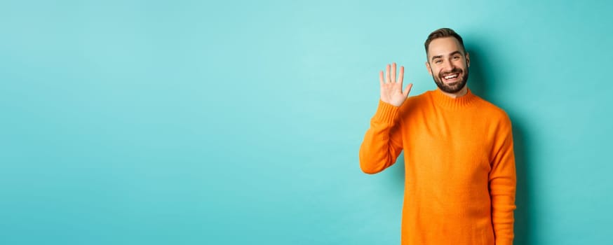 Photo of friendly young man saying hello, smiling and waiving hand, greeting you, standing in orange sweater over light blue background.
