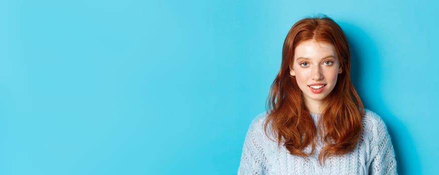 Close-up of young cute redhead girl smiling at camera, standing against blue background.