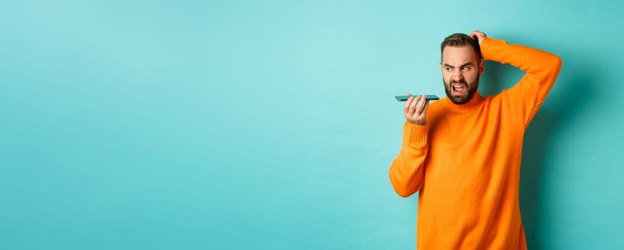 Confused man scratching head while talking on speakerphone, record voice message with indecisive face, standing in orange sweater over light blue background.