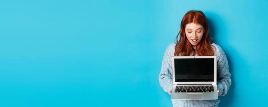 Amazed redhead girl staring at laptop screen and looking impressed, showing computer display, standing over blue background.