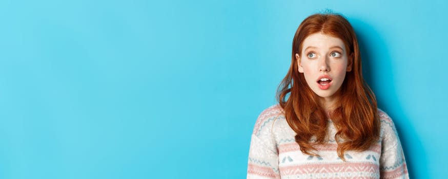 Close-up of impressed redhead girl staring left amazed, open mouth in awe and glancing at promo, standing in winter sweater against blue background.