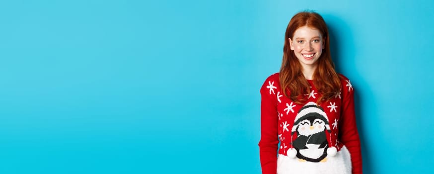Winter holidays and Christmas Eve concept. Cute smiling teenage girl with red hair, wearing funny xmas sweater, standing against blue background.