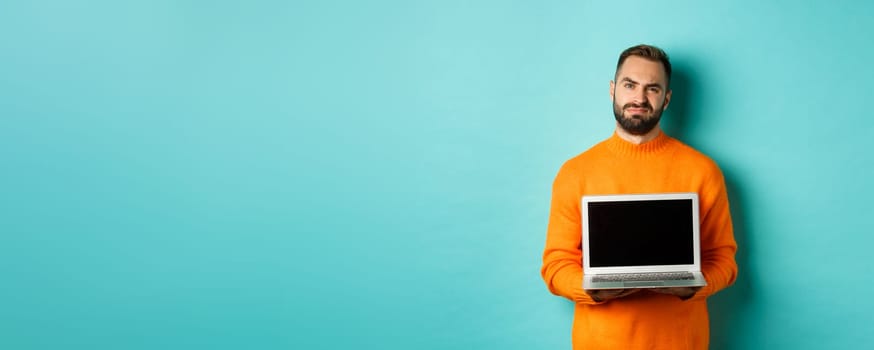 Handsome bearded man in orange sweater showing laptop screen, demonstrating promo, grimacing disappointed and upset standing over light blue background.