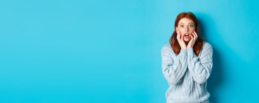 Startled redhead girl staring at something amazing, gasping in awe, standing in sweater against blue background. Copy space