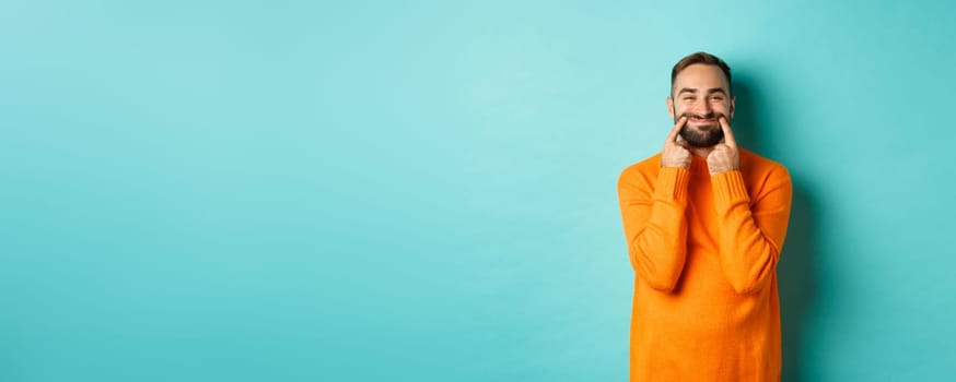 Image of bearded man stretching lips in happy smile, faking happiness, standing over light blue background. Copy space