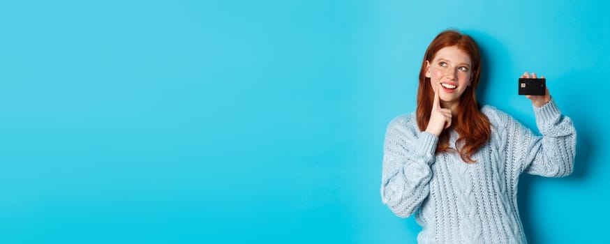 Image of thoughtful redhead girl thinking about shopping, showing credit card and pondering, standing over blue background.