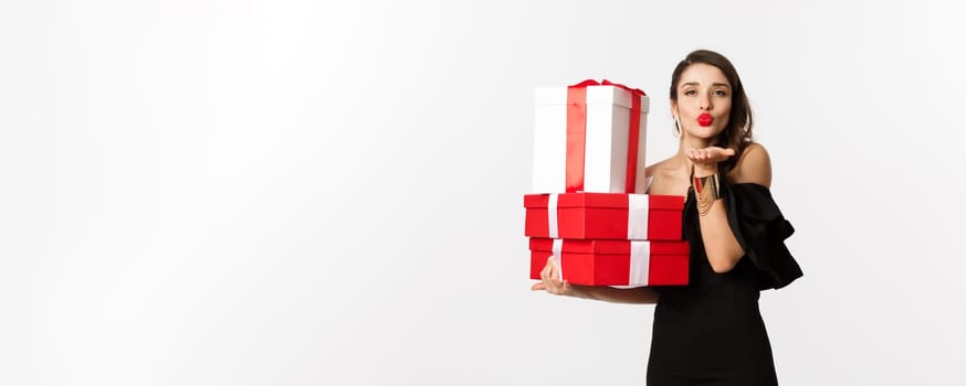 Celebration and christmas holidays concept. Pretty woman in elegant black dress holding presents, sending air kiss at camera, standing over white background.