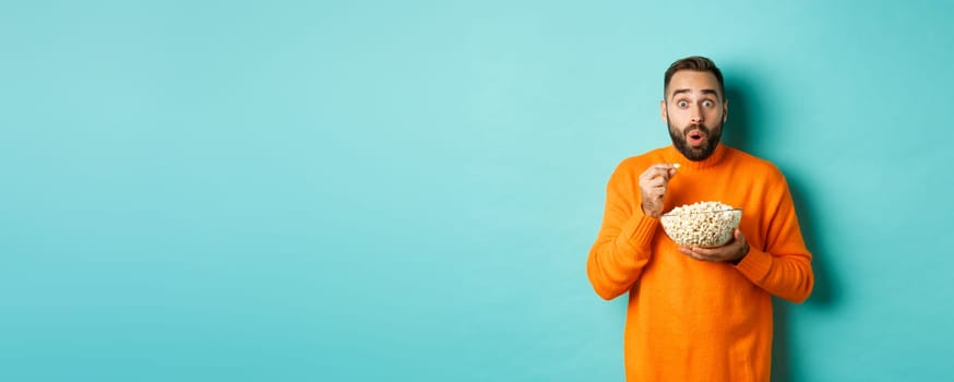 Excited young man watching interesting movie on tv screen, eating popcorn and looking amazed, blue background.