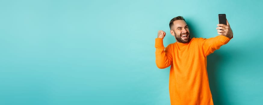 Handsome man video calling, looking happy and rejoicing of good news, making fist pump, communicating via mobile phone, standing over light blue background.