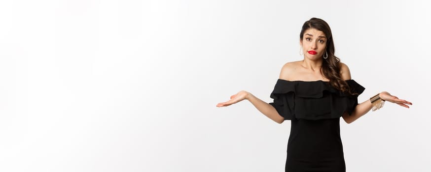 Fashion and beauty. Clueless attractive woman in black dress dont know, shrugging and smirking unaware, standing indecisive over white background.