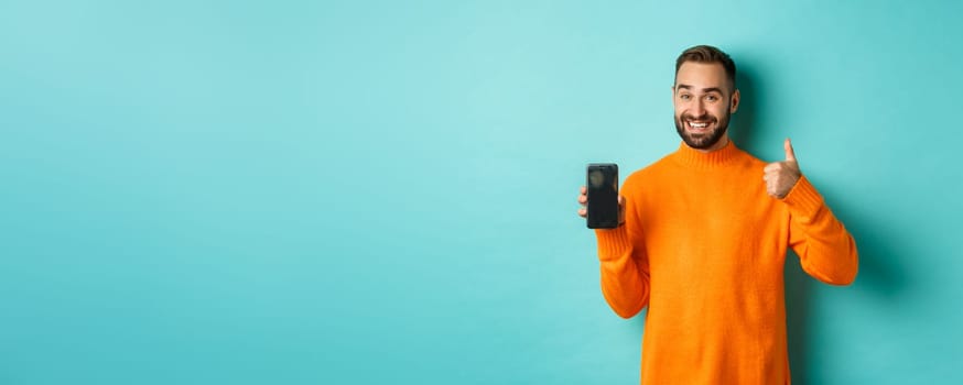 Attractive bearded man showing smartphone screen, thumbs up, recommending mobile app, standing satisfied over turquoise background.