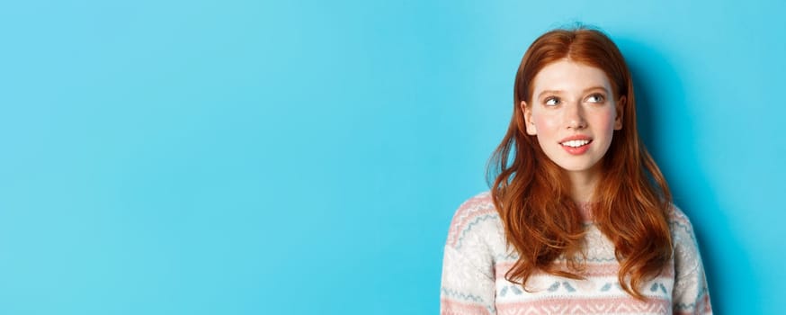 Close-up of dreamy redhead girl imaging something, staring at upper right corner and smiling, standing in winter sweater against blue background.