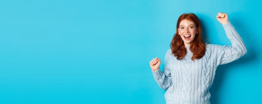 Cheerful redhead gil winning, celebrating victory, smiling and jumping from happiness, posing against blue background.