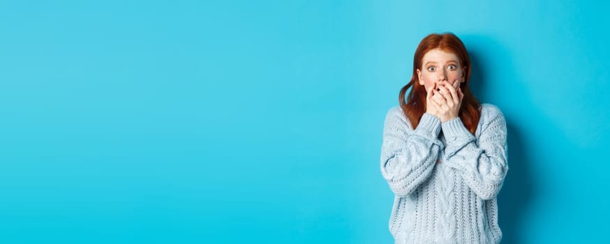 Shocked redhead girl in sweater, staring at camera scared and gasping, covering mouth with hands, standing against blue background.
