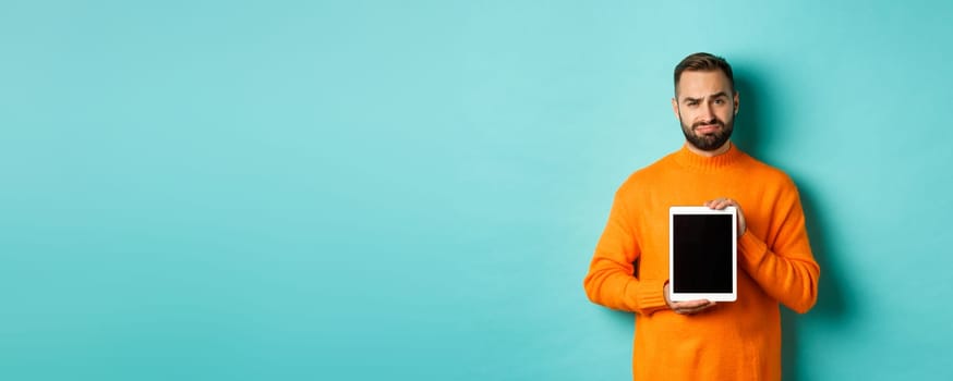 Technology. Skeptical and displeased guy showing digital tablet screen, frowning disappointed, standing over light blue background.