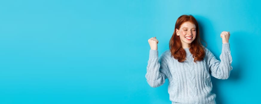 Satisfied redhead girl achieve goal and celebrating, making fist pump gesture and smiling with rejoice, triumphing of win, standing against blue background.