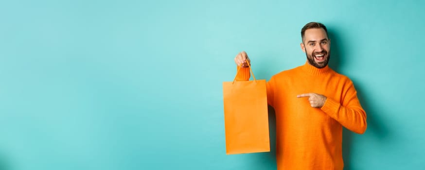 Satisfied male customer pointing at orange shopping bag, recommending store, smiling pleased, standing over turquoise background.