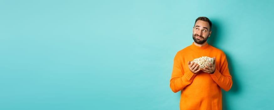 Handsome young man in orange sweater, looking thoughtful at upper left corner, holding popcorn, picking movie, blue background.