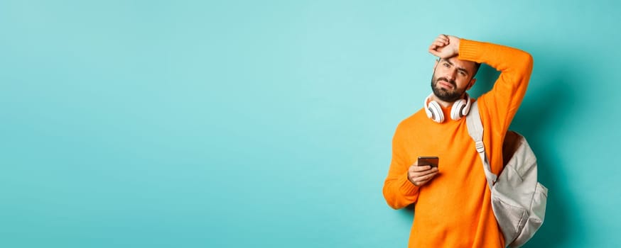 Tired guy with headphones and backpack, wiping sweat off forehead with exhausted face, using mobile phone, standing over turquoise background.