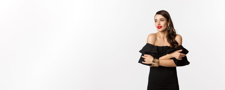 Beauty and fashion concept. Elegant and beautiful woman in black dress, makeup, embracing herself and looking away with sensual gaze, standing over white background.