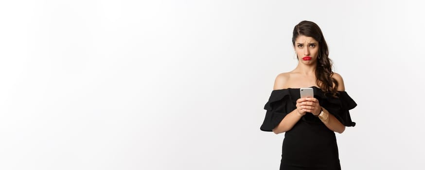 Sad and gloomy woman in black dress, sulking upset, using mobile phone and feeling disappointed, standing over white background.