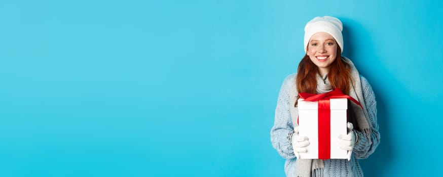 Winter holidays and Christmas sales concept. Cute redhead teeanage girl in beanie, sewater and scarf holding gift in wrapped box, smiling, wishing merry xmas, standing over blue background.