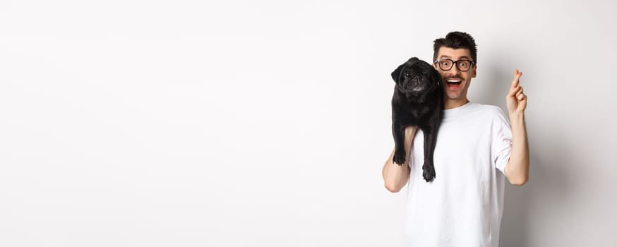 Hopeful smiling dog owner making a wish, holding cute black pug on shoulder and cross fingers for good luck, white background.