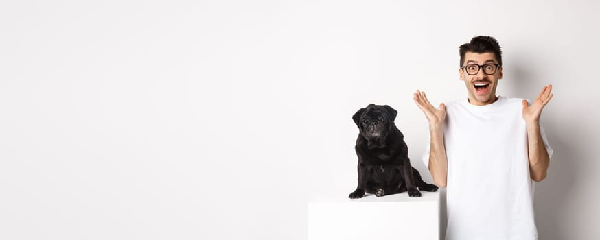 Cheerful young man in glasses standing with his pet, rejoicing and staring at camera amused, hear great news, standing with pug over white background.