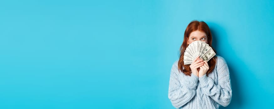 Thoughtful cute girl with red hair dreaming about shopping, holding dollars and looking at upper left corner logo, standing over blue background.