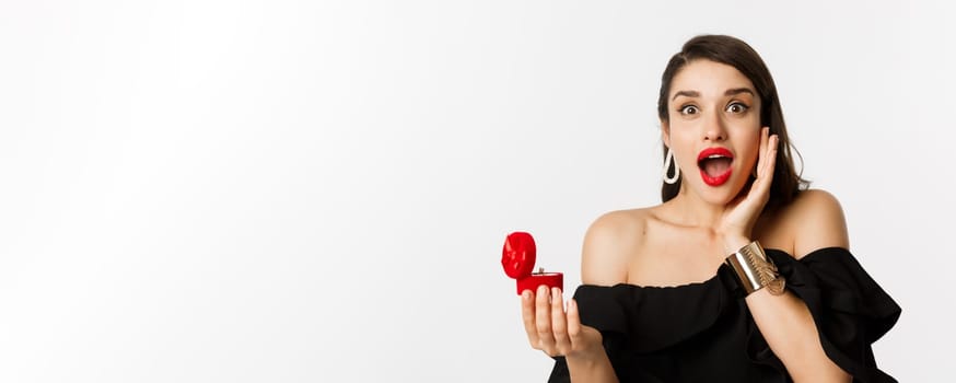 Close-up of attractive young woman with red lipstick, makeup on, looking amazed after receiving marriage proposal, holding engagement ring, standing over white background.