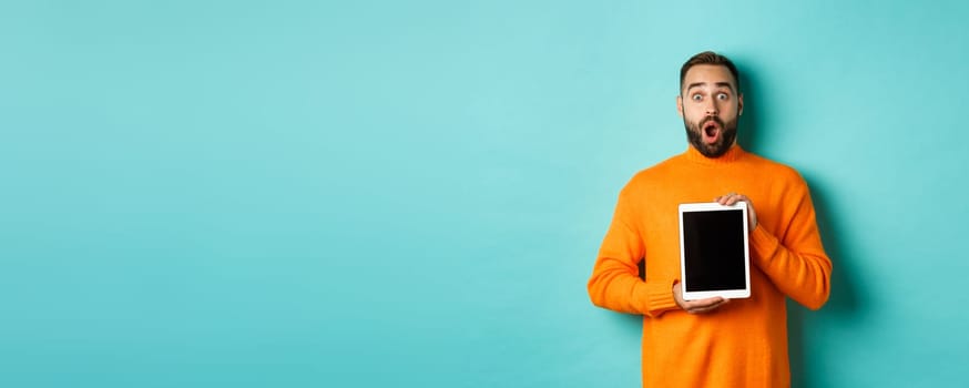 Technology. Surprised bearded man drop jaw, staring at camera and showing digital tablet screen, standing againt light blue background.