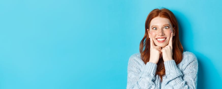 Close-up of funny redhead teen girl making faces, squinting and squeezing cheeks, standing against blue background.