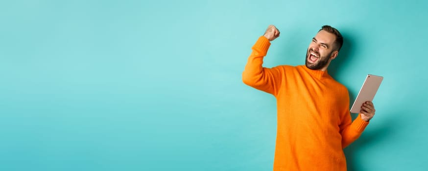 Cheerful winning man holding digital tablet, rejoicing and celebrating victory in game, making fist pump gesture, standing over light blue background.