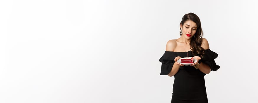 Party and celebration. Cute young woman with red lips, wearing black dress, holding birthday cake with candle and looking dreamy, making wish, white background.