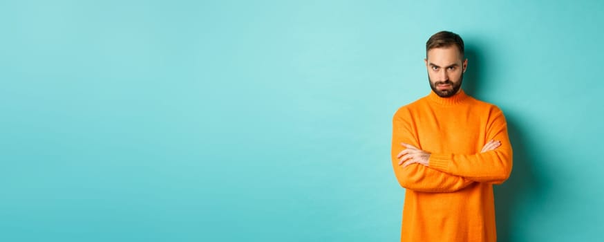 Offended man looking angry at you, cross arms on chest and stare mad, standing in orange sweater against turquoise background.