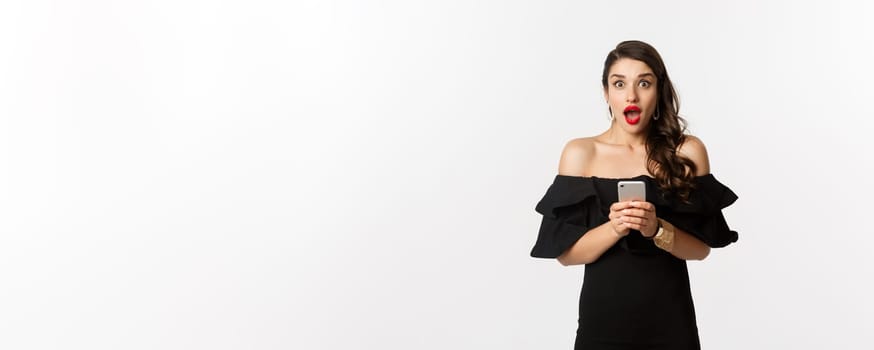 Happy young glamour woman in black dress, holding mobile phone and looking surprised, standing over white background.