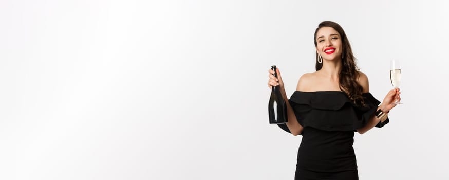 Celebration and party concept. Stylish brunette woman in glamour dress holding bottle and glass of champagne, smiling pleased, standing over white background.
