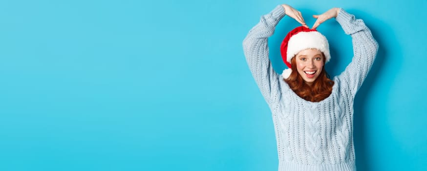 Winter holidays and Christmas Eve concept. Cute redhead teen girl in santa hat and sweater, making heart sign and smiling, wishing merry xmas, standing over blue background.