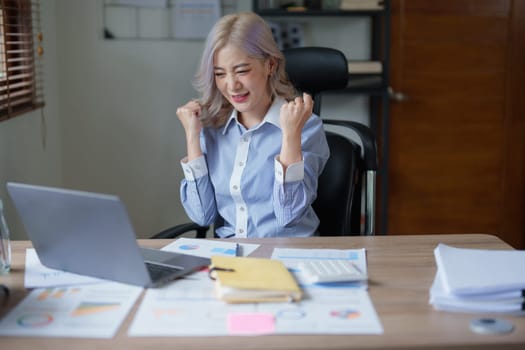 Portrait of a woman business owner showing a happy smiling face as he has successfully invested her business using computers and financial budget documents at work.