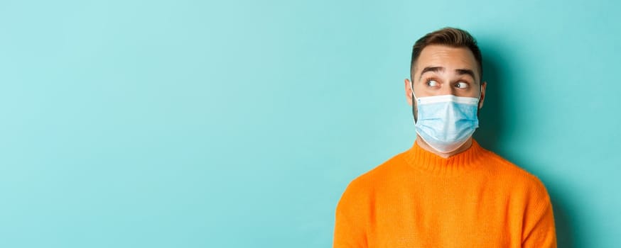 Covid-19, social distancing and quarantine concept. Close-up of young male model in medical mask looking left curious, staring at promo copy space, light blue background.