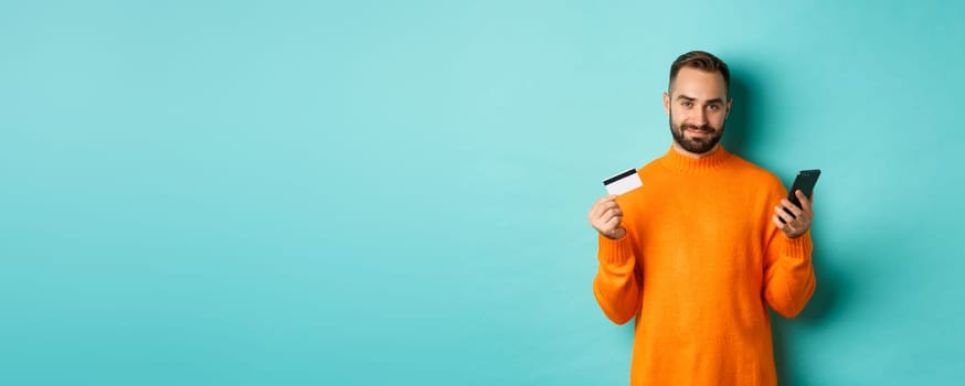 Online shopping. Handsome caucasian man in orange sweater, using credit card and mobile phone, standing over light blue background.