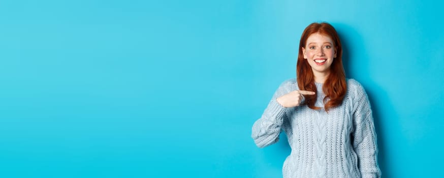 Beautiful redhead girl pointing at herself and smiling happy, being chosen, standing in sweater against blue background.