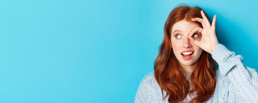 Headshot of pretty redhead girl in sweater, looking left at promo with okay sign over eye, standing against blue background.