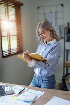 Portrait of a young Asian woman showing a smiling face as she using notebook, computer and financial documents on her desk in the early morning hours.