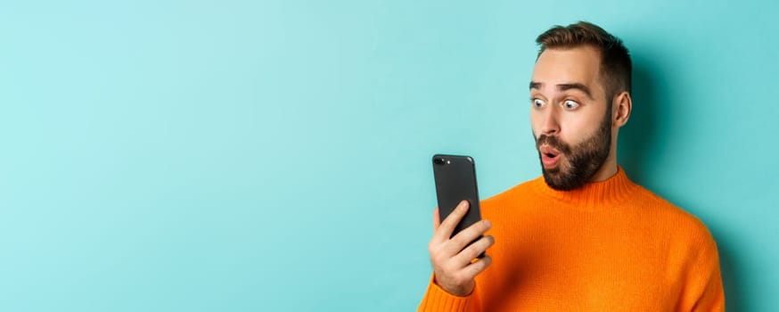 Close-up of caucasian man staring at phone screen with surprised face, wearing orange sweater, light blue background.