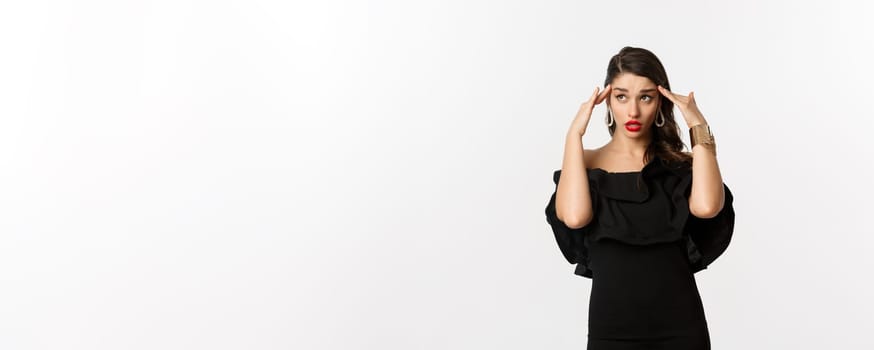 Fashion and beauty. Annoyed and tired woman in black dress, touching head and roll eyes bothered, standing distressed against white background.