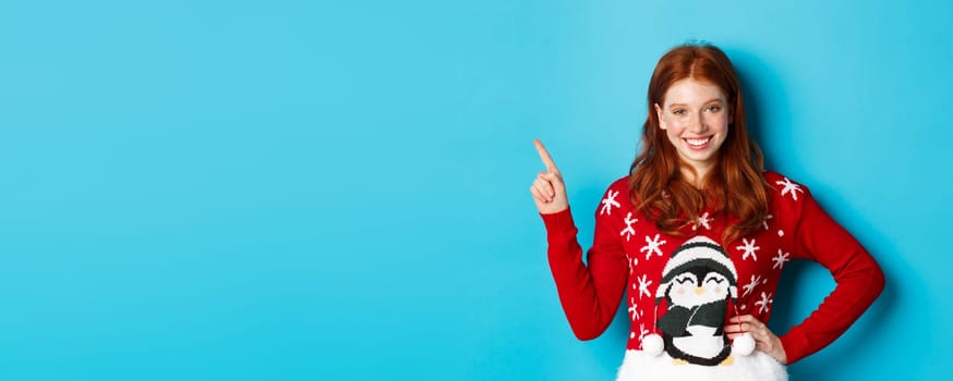 Winter holidays and Christmas Eve concept. Cute teenage girl with red wavy hair, pointing upper left corner and smiling at camera, standing over blue background.