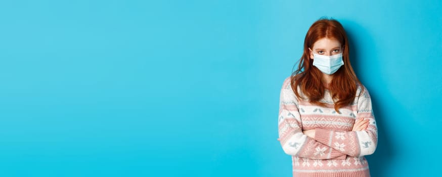 Winter, covid-19 and pandemic concept. Skeptical redhead girl in medical mask, cross arms on chest and stare angry at camera, standing over blue background.